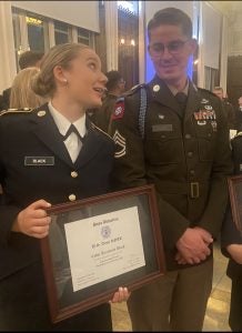 Two Army cadets in dress uniform look at each other while one carries an award