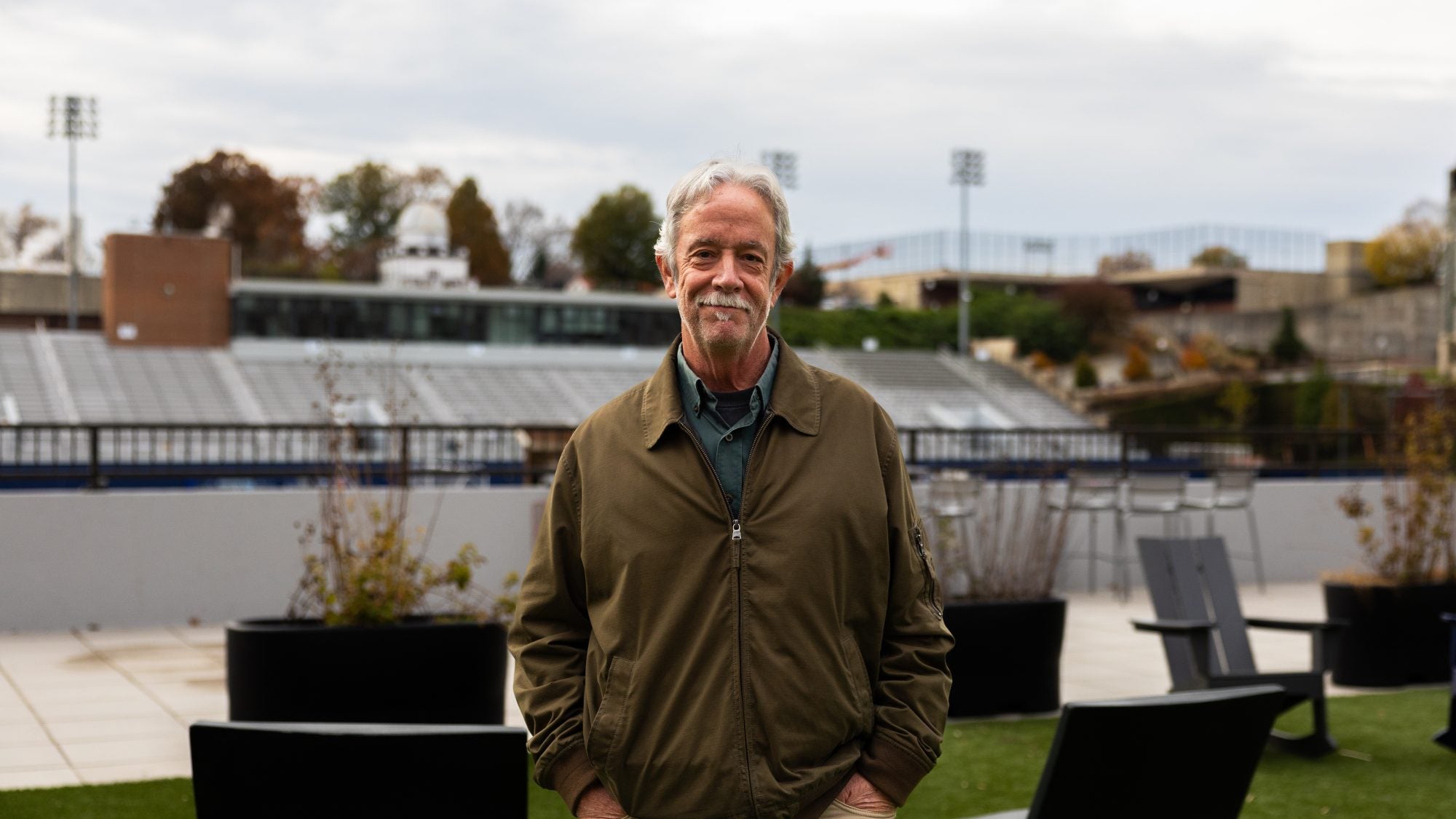 A man with gray hair and a mustache stands in front of a football field. He wears an olive jacket.