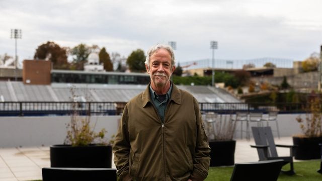 A man with gray hair and a mustache stands in front of a football field. He wears an olive jacket.