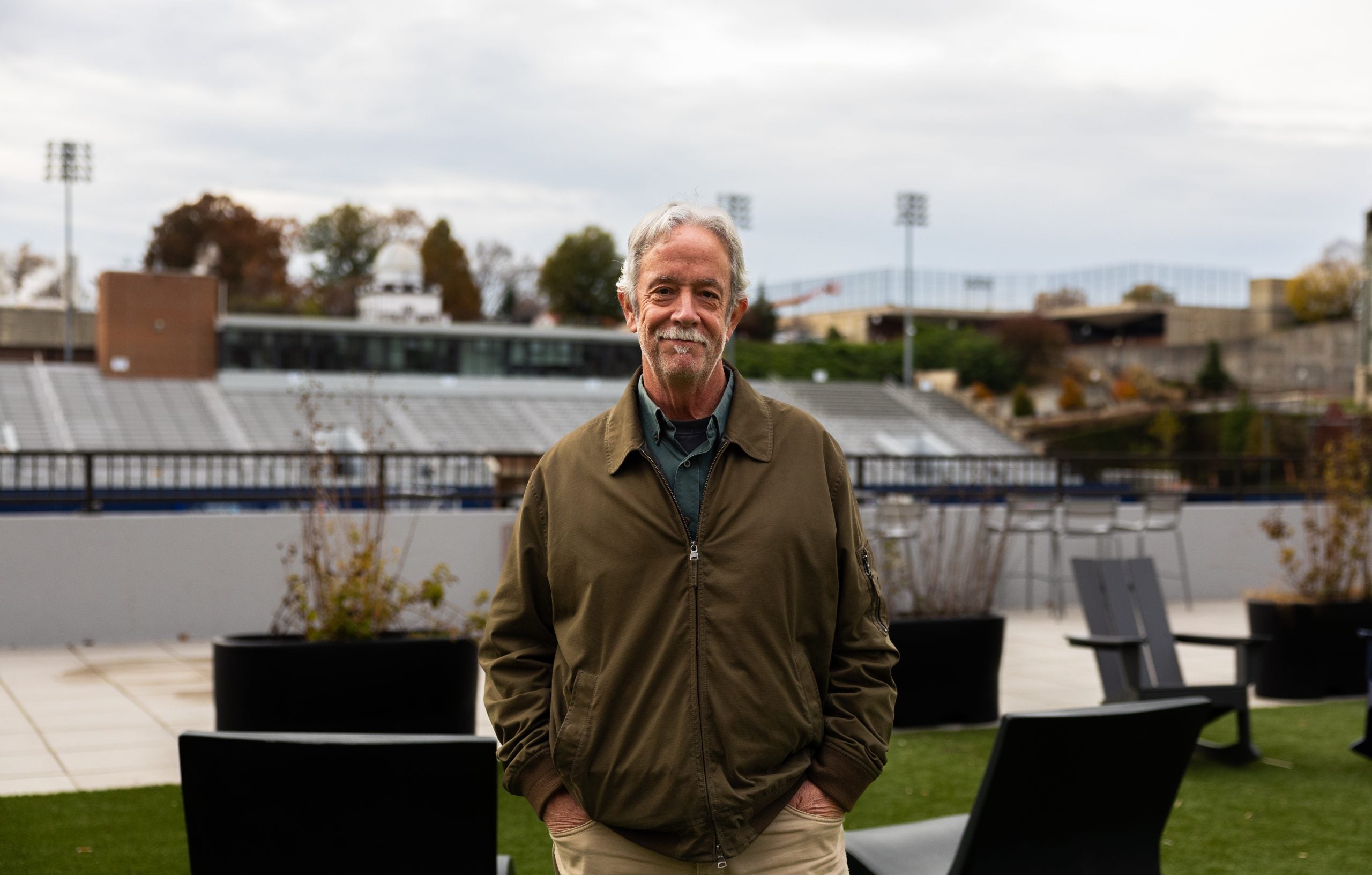 A man with gray hair and a mustache stands in front of a football field. He wears an olive jacket.