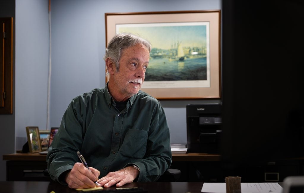 A man with gray hair and a mustache looks at his computer while writing in an office setting.