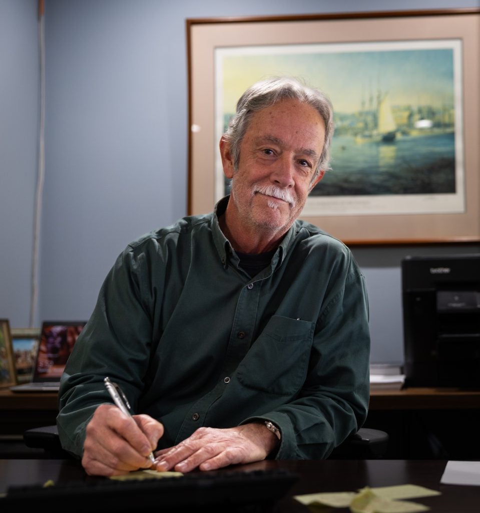 A man with a mustache smiles as he writes at a desk in an office.