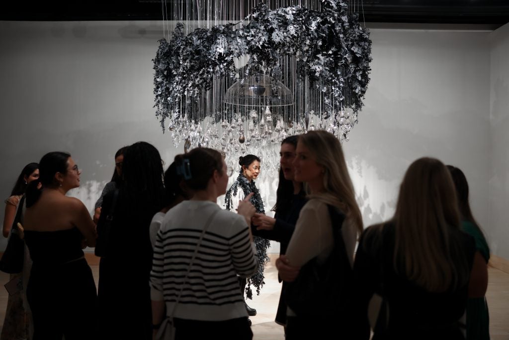 A group of people talk at an art gallery opening. In the background, an artist stands under her interactive art, in which hand-blown glass dangles from the ceiling with a clear white dome in the middle.