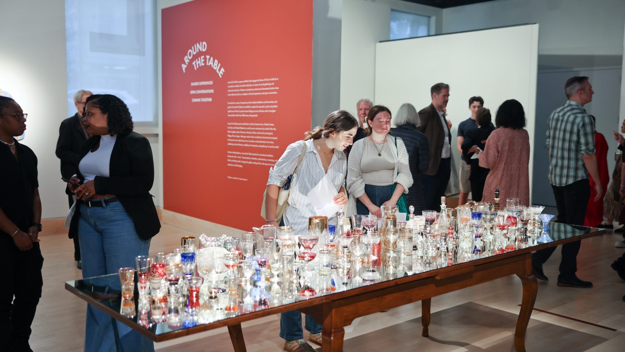 Two women peer at a table with glasses on it as part of a display in an art gallery.