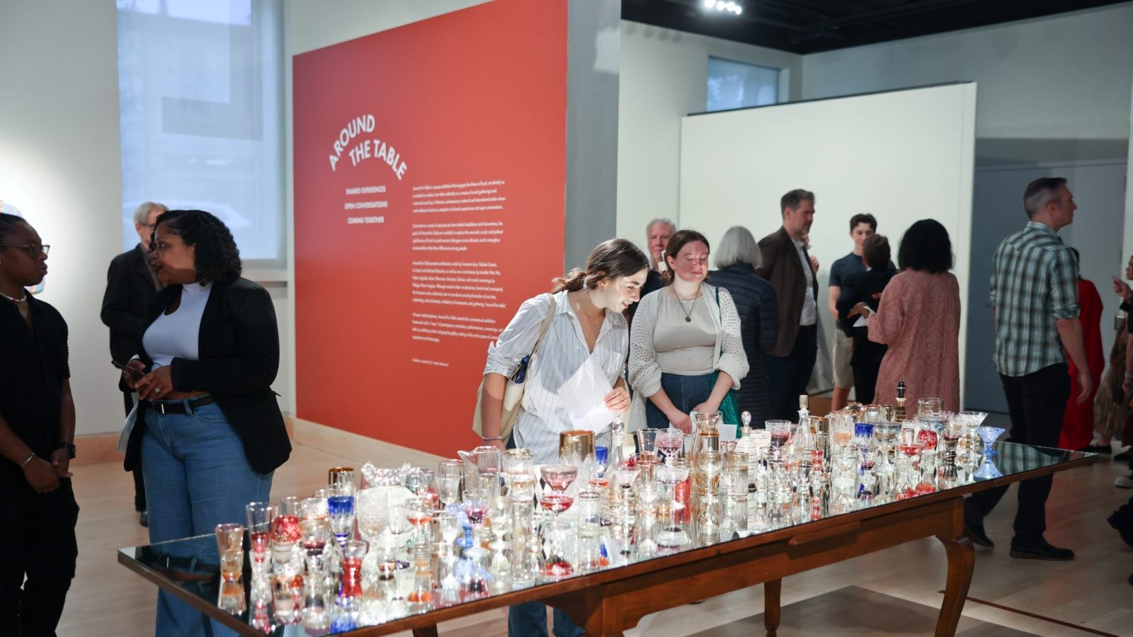 Two women peer at a table with glasses on it as part of a display in an art gallery.