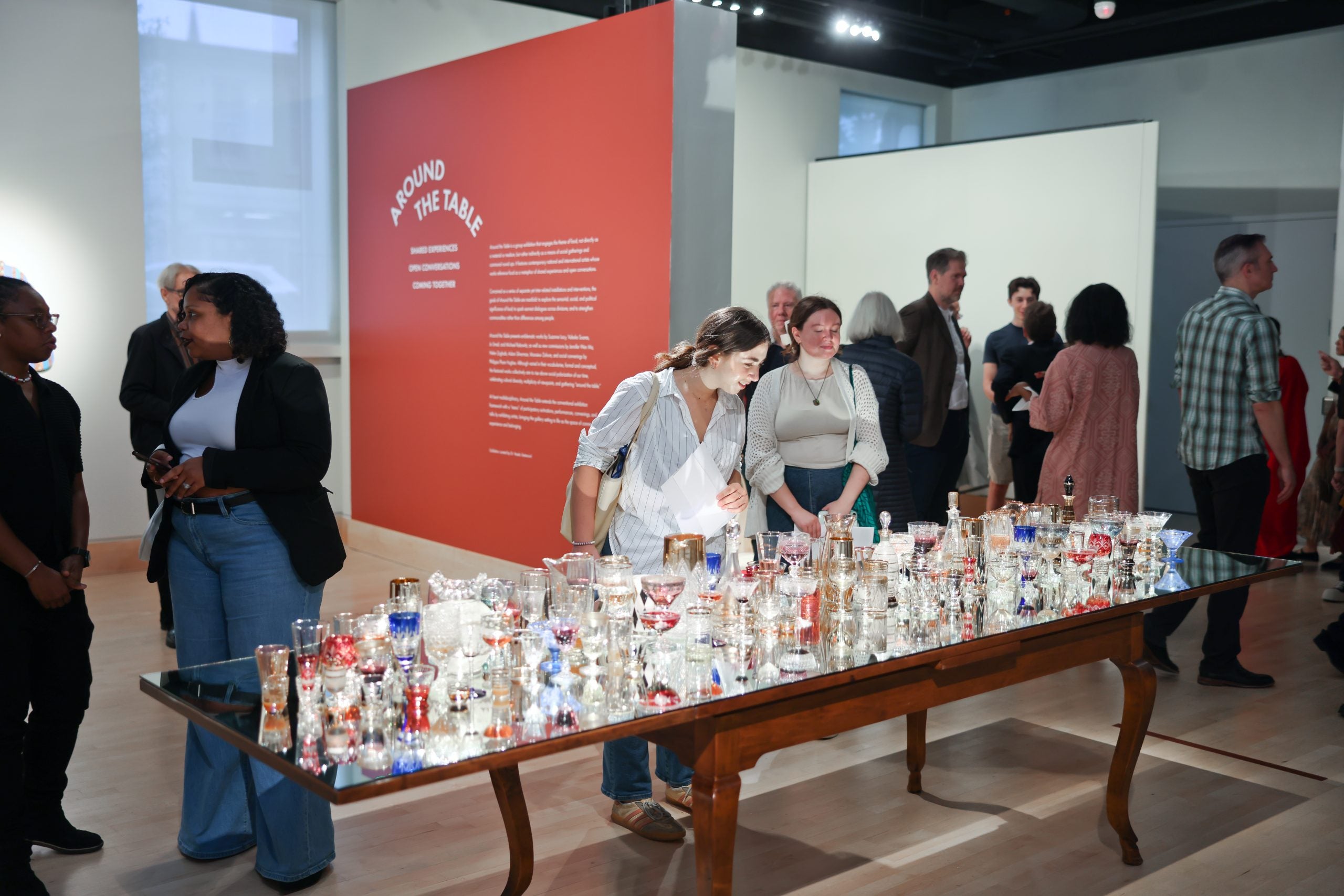 Two women peer at a table with glasses on it as part of a display in an art gallery.