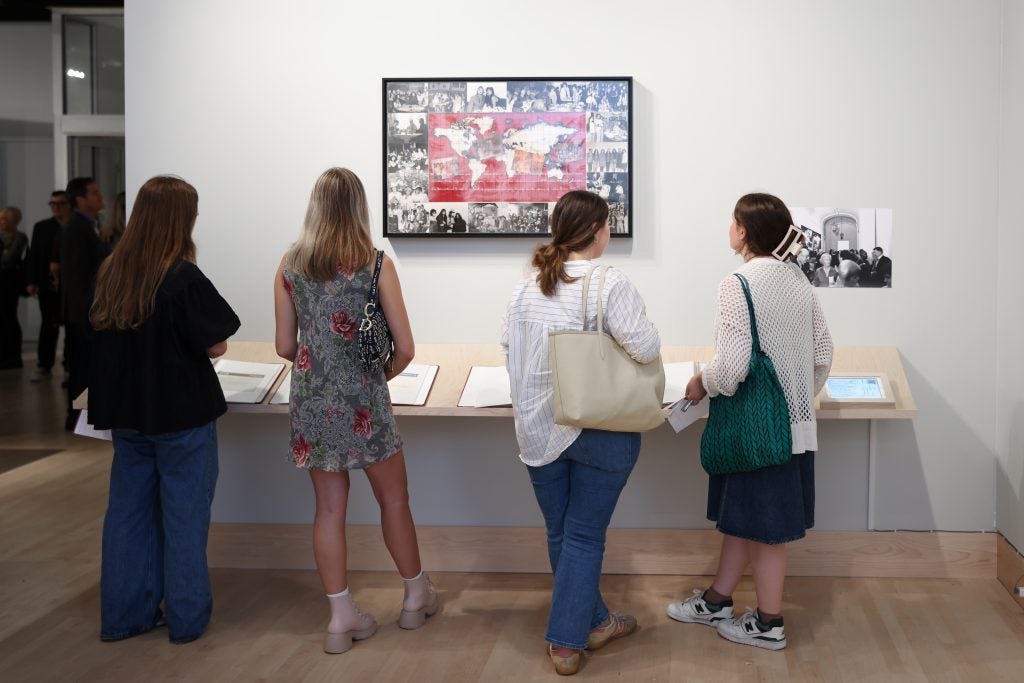 A group of women look at an interactive exhibit at a gallery. Above them is a black and white collage with red in the center.