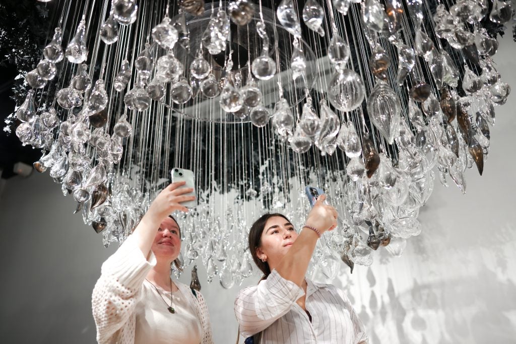 Two women stand underneath a series of hand-blown glass that dangles from the ceiling. They take pictures of the artwork.