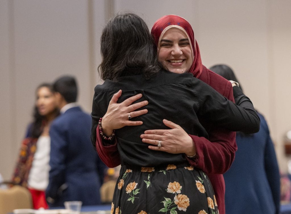 Two females hug each other at an event. One of the students facing the camera and smiling wears a red hijab.