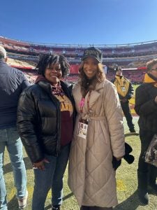 Two women stand on a football field with a stadium behind them on a sunny day.