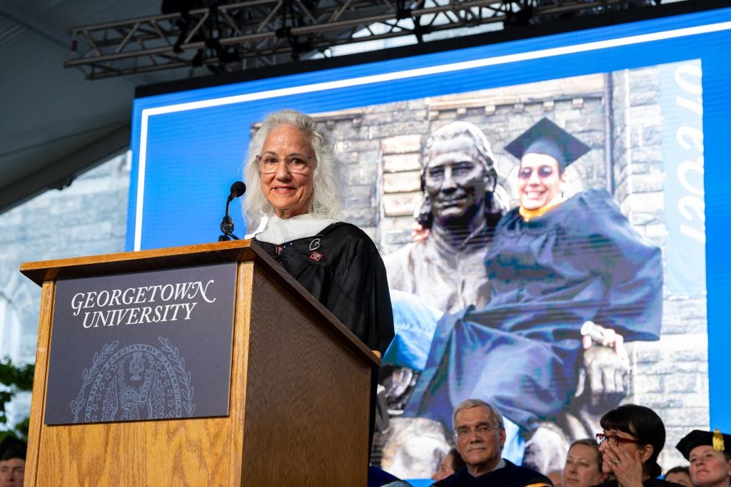 Debra Tice speaking at a podium at GU commencement in a commencement gown with a photo of her son Austin behind her.