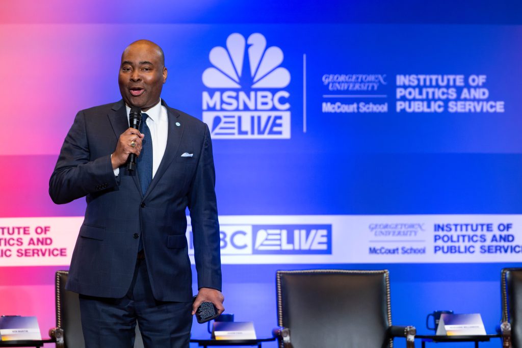 A Black man in a blue suit and tie speaks on stage with a microphone and a MSNBC background behind him