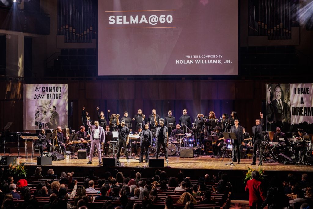 A group of performers stand with their fists in the air on a stage. Behind them are the words "Selma@60"