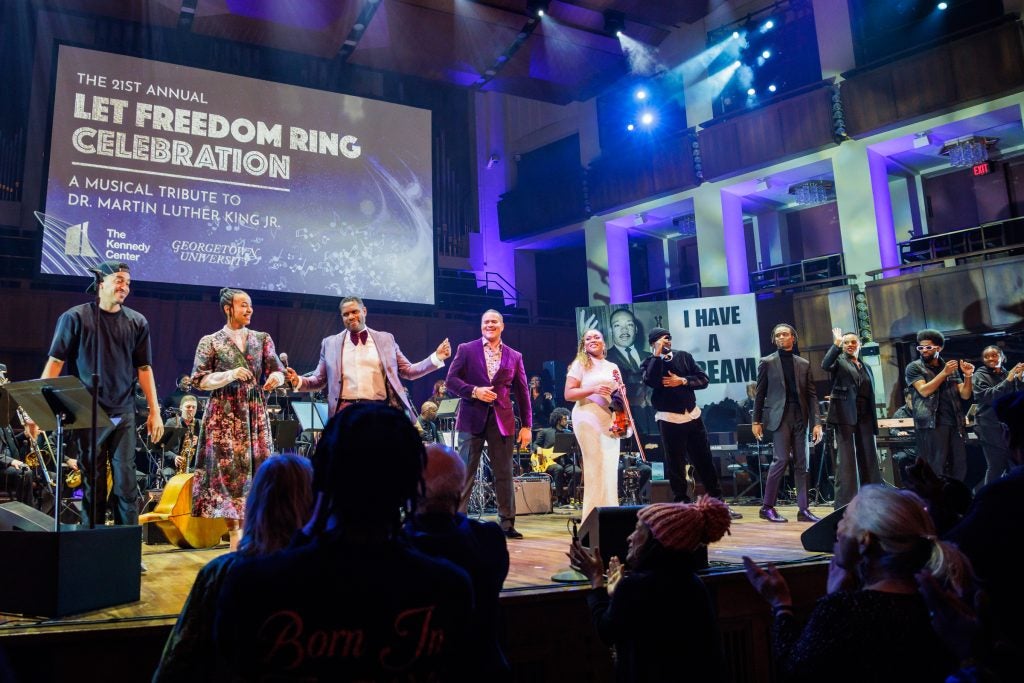 A group of performs sing and dance onstage. Behind them on a screen says, "Let Freedom Ring Celebration."