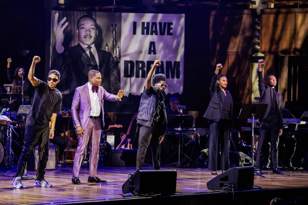 A group of performers dressed in black raise one fist in the air. The performance's director stands in the middle in a lavender suit.