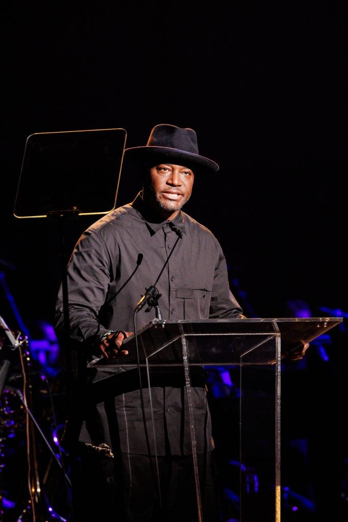 Actor Taye Diggs introduces a performance while standing on a stage behind a podium. He is dressed in all black with a black hat.