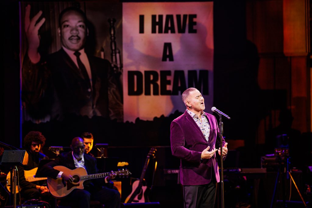 Christopher Jackson sings into a microphone while wearing a purple blazer.