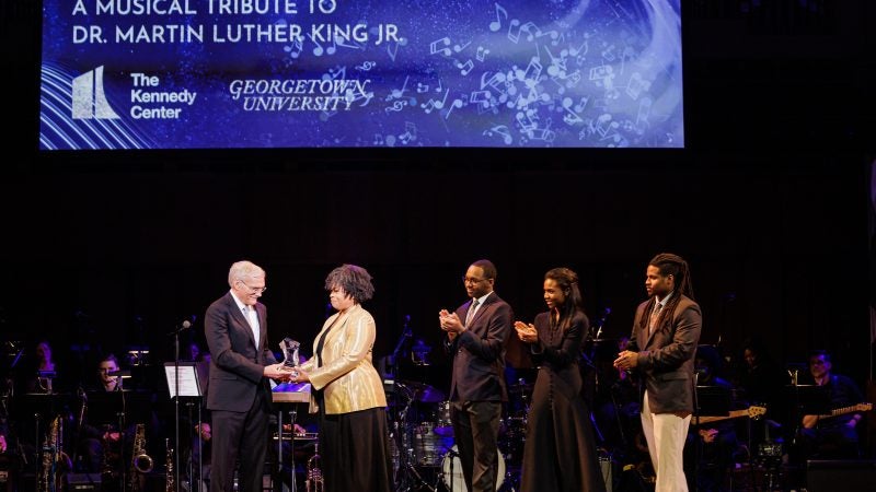 A woman in a beige blazer accepts an award from Georgetown&#039;s interim president on a stage. Next to her, three people clap.