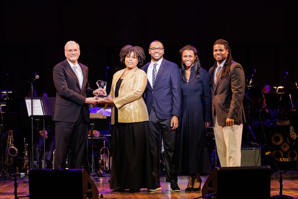 A woman in a shiny beige blazer accepts an award onstage next to four others.