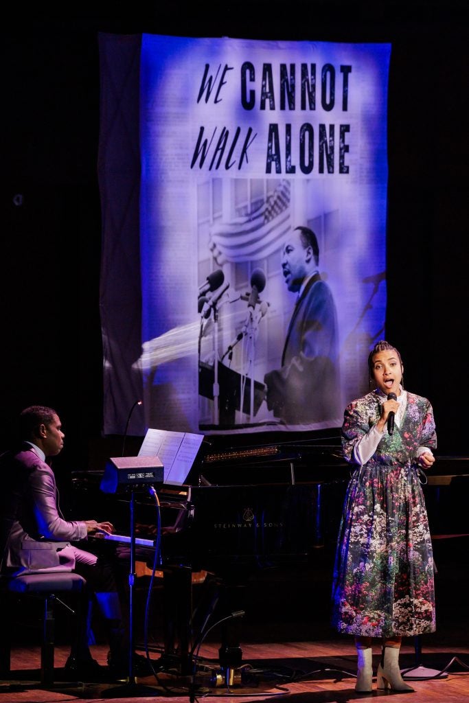 Esperanza Spalding sings into a microphone on a stage. Next to her, a musician plays the piano.