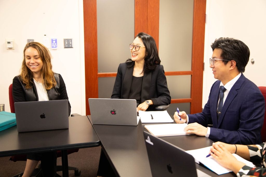 Two women and a male student smile and sit around an L-shaped table with computers and papers.