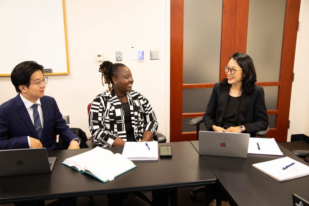 Two students smile at a professor as they sit around an L-shaped table.