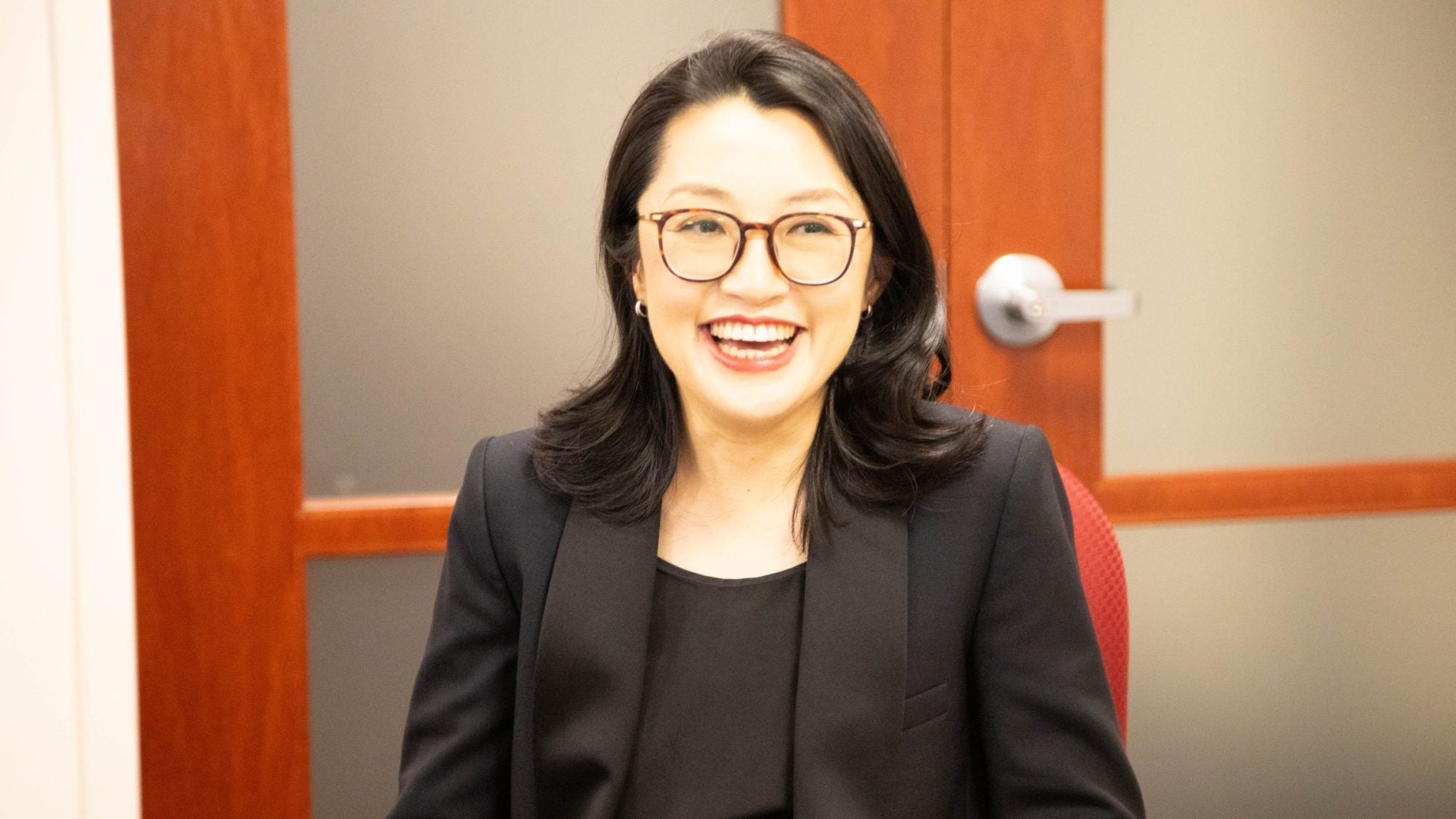 A woman with black hair, glasses and a black blazer smiles while sitting behind a table with a laptop in front of her.