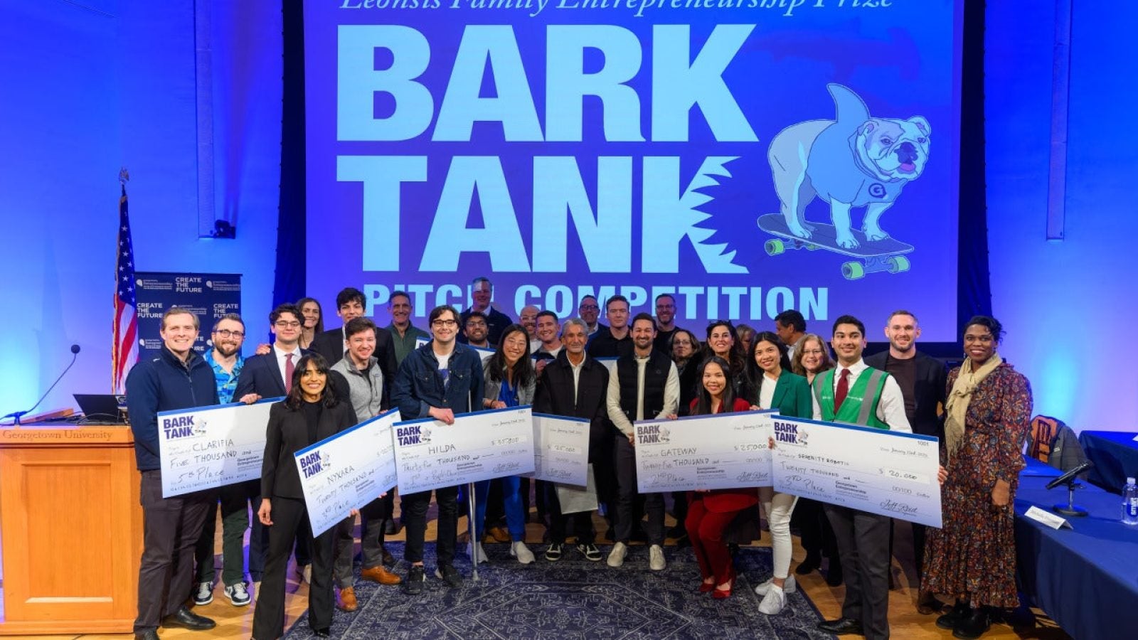 A group of students hold up giant checks in front of a blue screen that says &quot;Bark Tank Pitch Competition&quot;