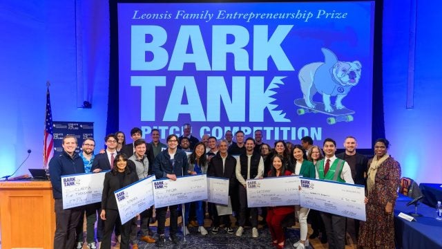 A group of students hold up giant checks in front of a blue screen that says &quot;Bark Tank Pitch Competition&quot;