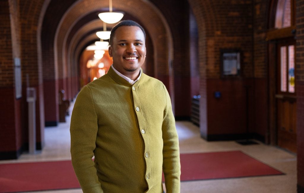 A man in a light green sweater smiles in a hallway.