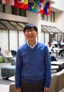 A man in a blue sweater and glasses laughs in an atrium.