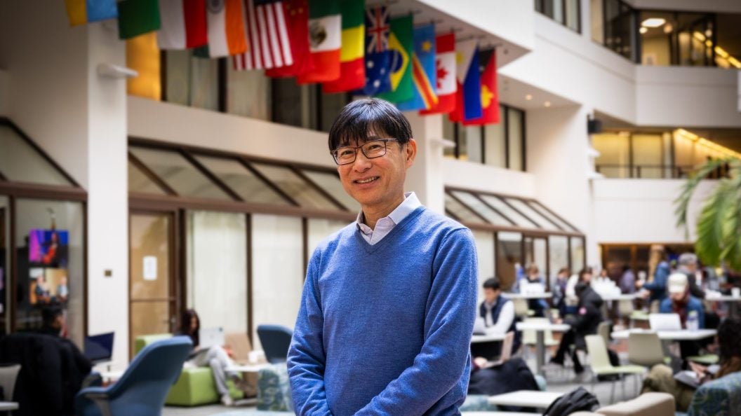 A man in a blue sweater smiles in front of hanging flags in an atrium.