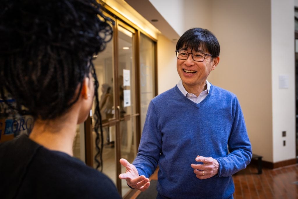 A man in a blue sweater and glasses smiles at a student.