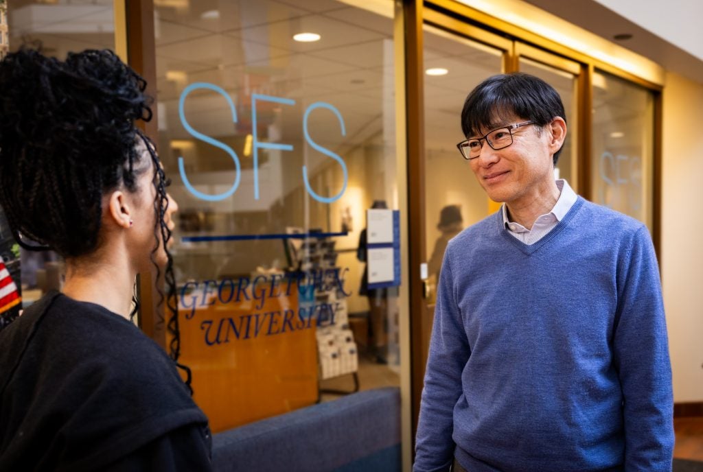 A man in a blue sweater and glasses listens to a student.