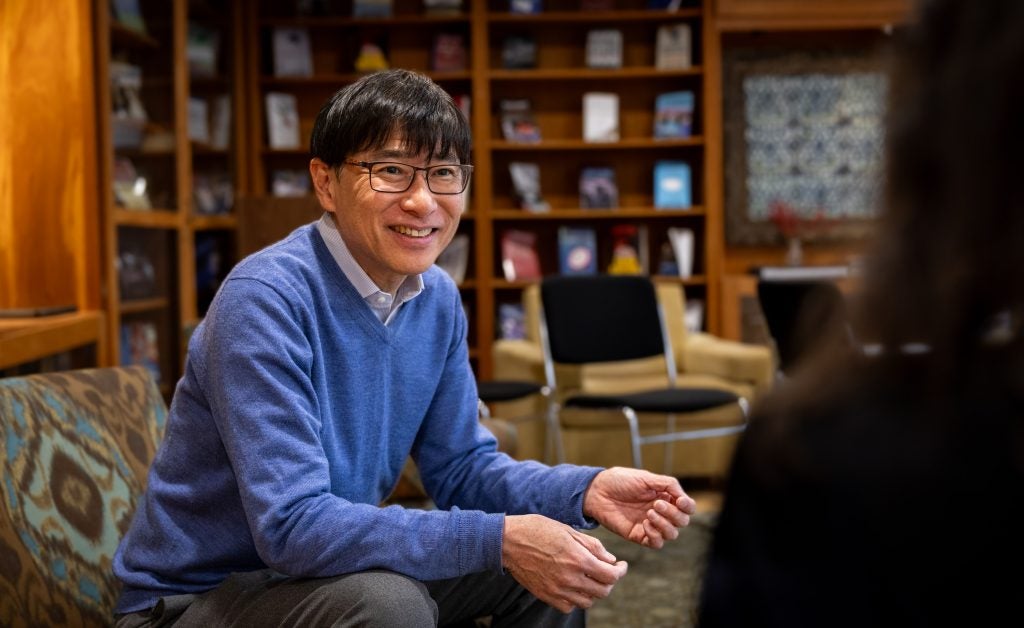 A man in a blue sweater and glasses sits and smiles while talking with a student.