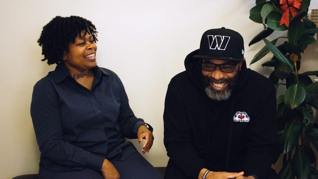 A man and woman laugh together. The man smiles while wearing a black sweatshirt and hat and the woman wears a navy blue button-down blouse.