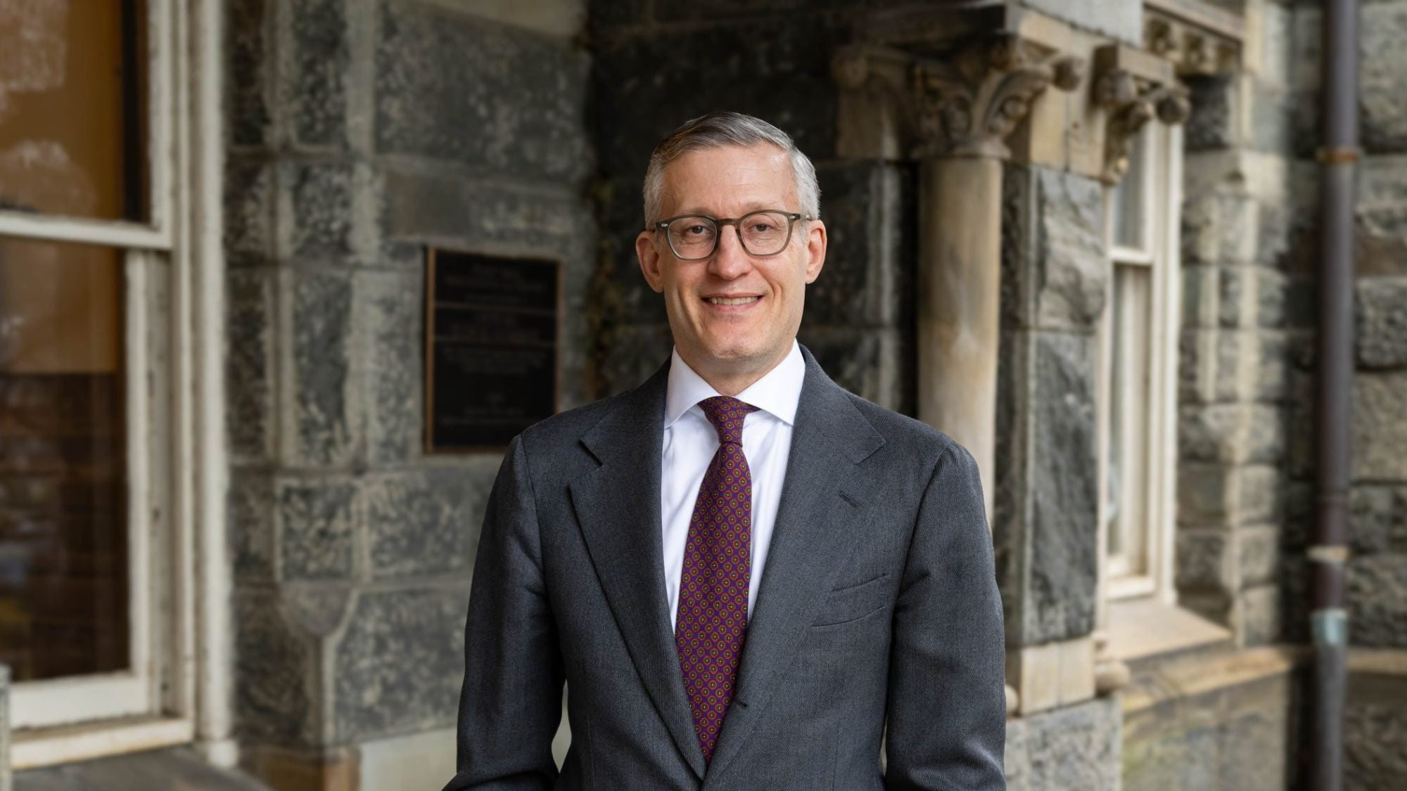 David Edelstein in a gray suit and dark red tie with hands in his pocket against Healy Hall background