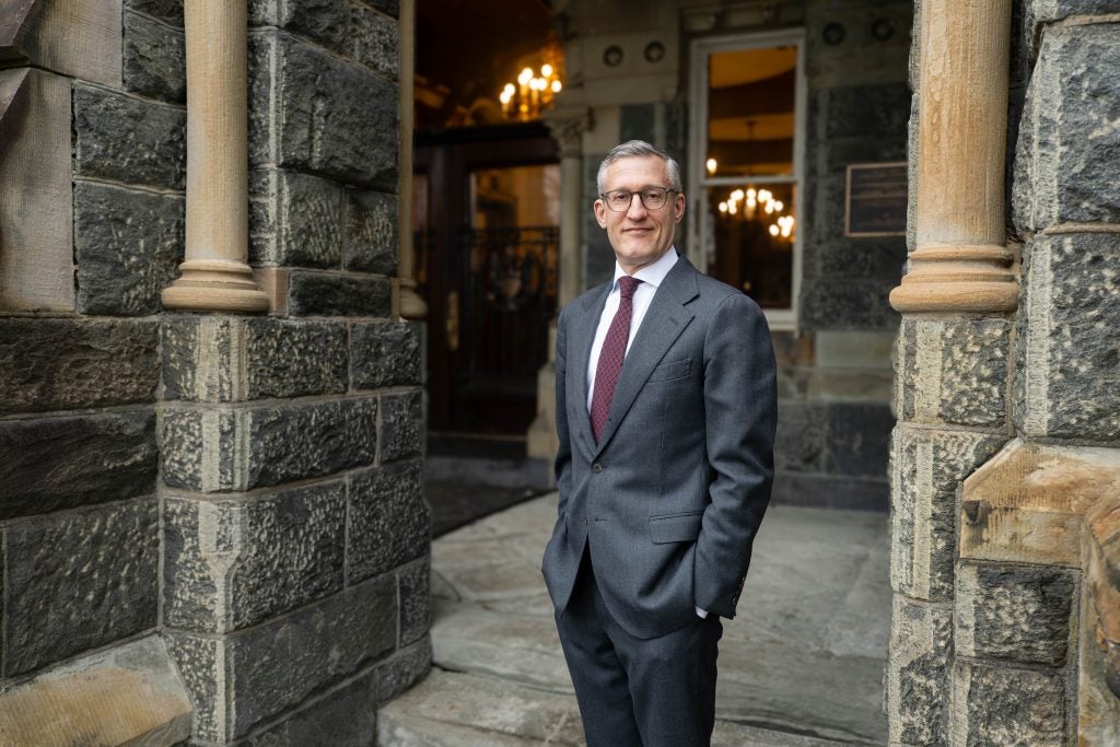 David Edelstein in a gray suit and dark red tie with hands in his pocket against Healy Hall background