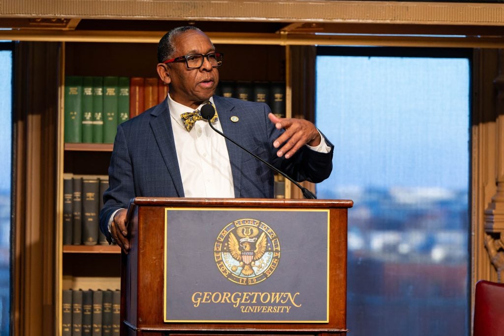 The president of Xavier University of Louisiana gestures with one hand while speaking behind a podium with a bookcase behind him.