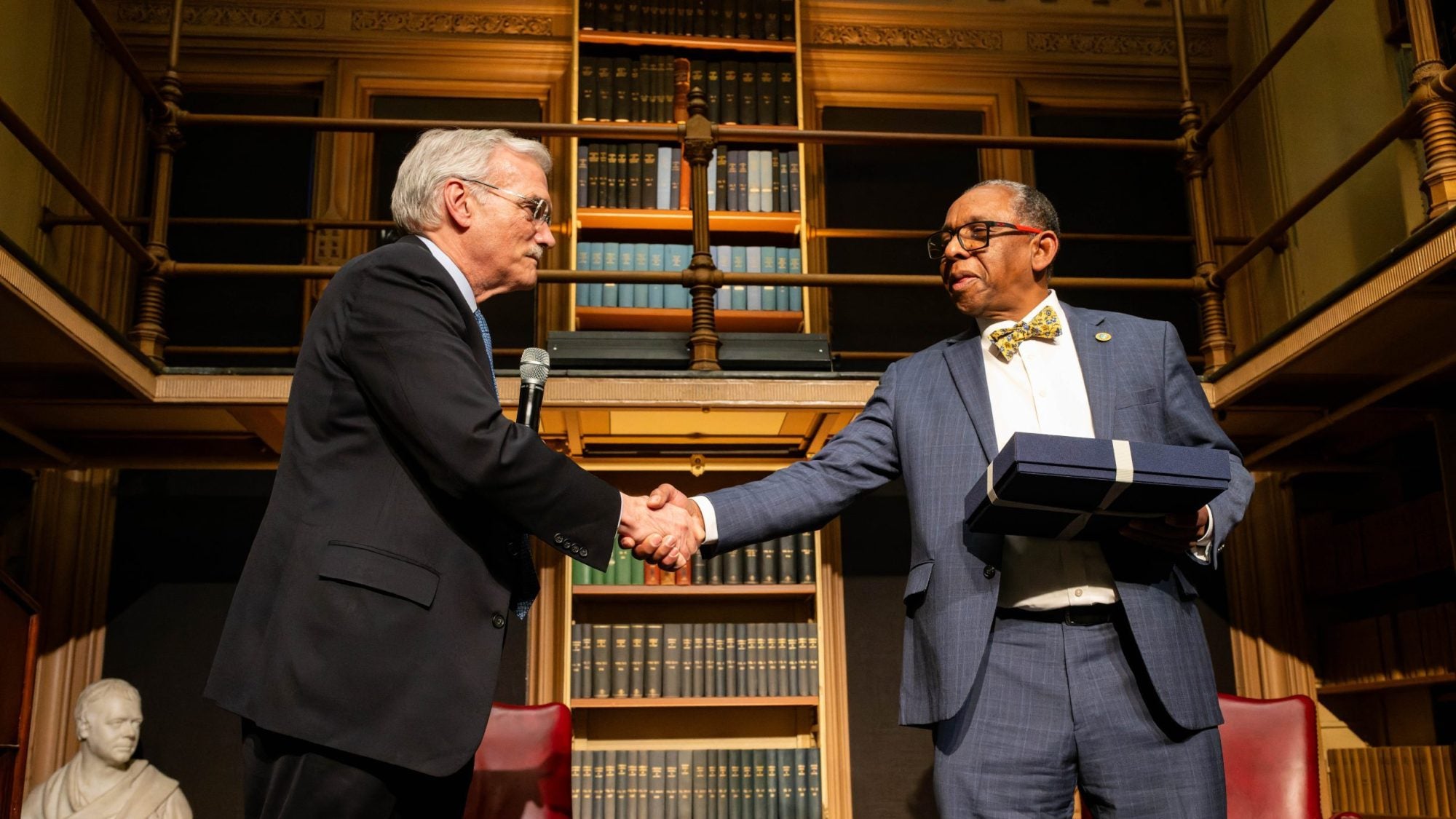 Two men shake hands on a stage with a bookshelf behind them.