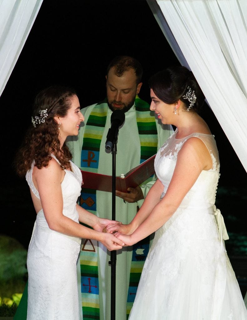 Two women wearing white dresses hold hands during their wedding ceremony.