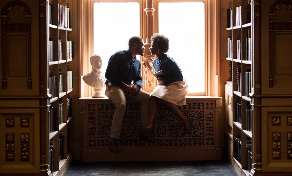 A couple sits on a ledge in a library and leans toward each other.