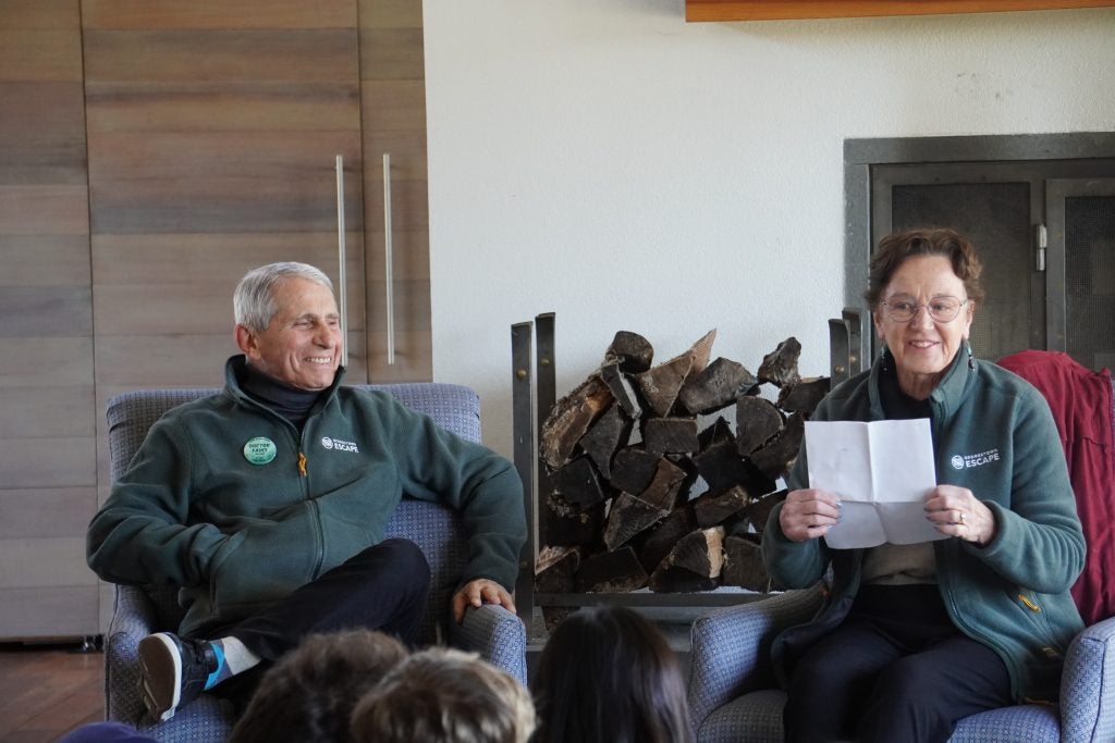 Dr. Anthony Fauci and his wife Christine Grady sit on chairs in front of a fireplace in a retreat center.