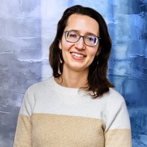 A woman with brown hair and glasses smiles in front of a textured blue background.