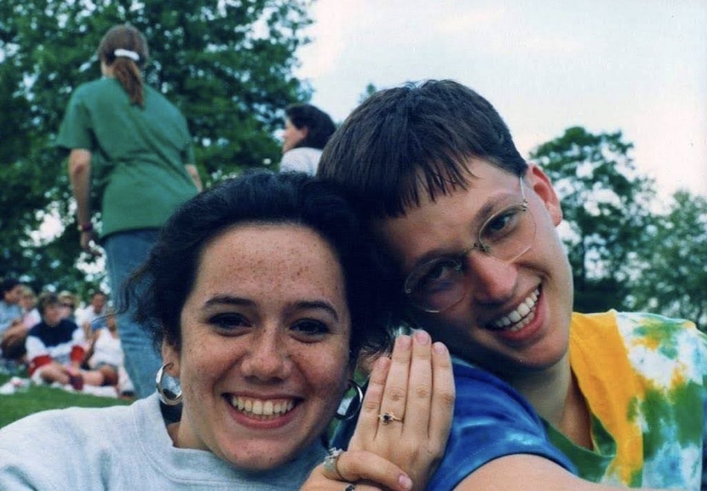 A woman holds up her engagement ring next to a man wearing a tie dye shirt.