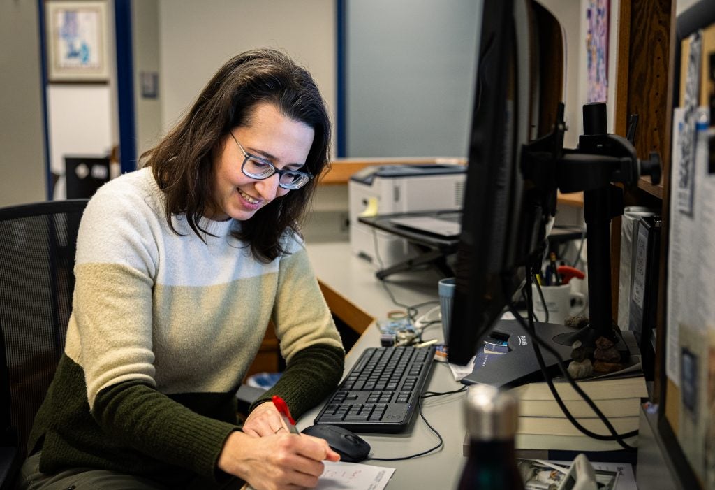 A woman smiles while writing on a pad of paper at her office desk.