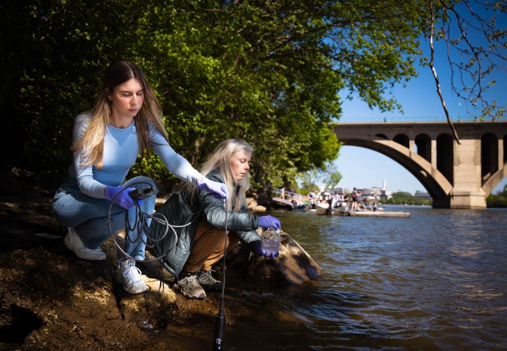 A student and a professor take water samples on the side of a river.