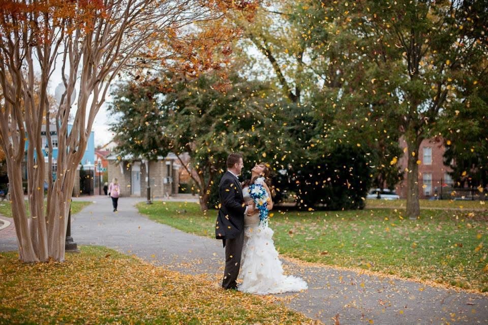 A bride and groom smile as orange leaves fall upon them.