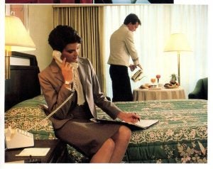 A woman is on the phone while sitting on a hotel bed while a man serves breakfast on a table by the window