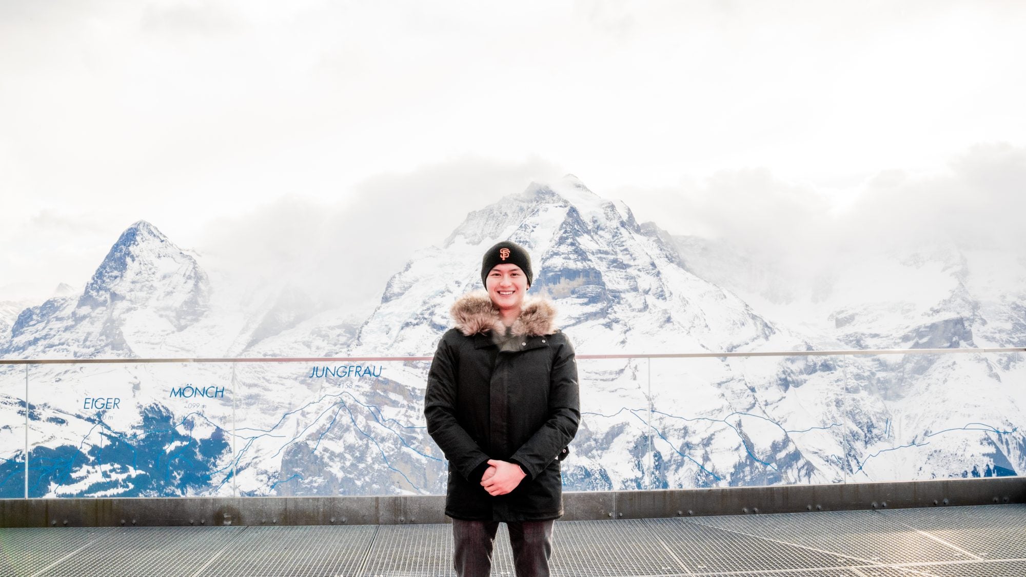 A young Asian man in a black coat with the Swiss Alps behind him